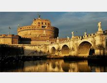 Veduta di Castel Sant’Angelo con la sua struttura cilindrica in muratura e, in primo piano, il Ponte Sant’Angelo in pietra con arcate e statue lungo il parapetto. Il ponte attraversa il fiume, che riflette le pareti illuminate e il cielo.