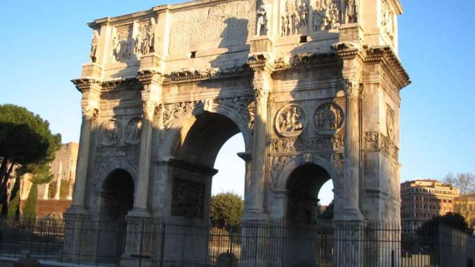 Arco monumentale romano in pietra con tre grandi aperture e ricchi bassorilievi decorativi sulle facciate. L’arco è circondato da una recinzione metallica, con alberi e edifici sullo sfondo sotto un cielo sereno.