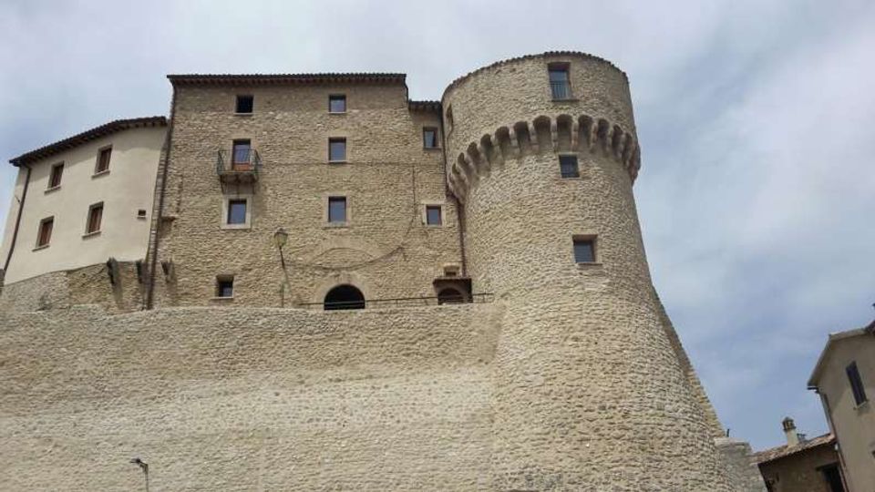 Castello in pietra con alta cinta muraria e torre cilindrica merlata, affacciato su un cielo nuvoloso. La struttura presenta facciate in muratura e intonaco con piccole finestre e balconcini, visibile da una prospettiva dal basso.