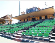 Gradinate di uno stadio con numerosi sedili verdi in file su livelli, delimitate da ringhiere metalliche. Sullo sfondo si vede la tribuna coperta con edificio in muratura gialla, finestre e struttura superiore vetrata.