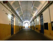 Lungo corridoio interno con soffitto a volta e archi ripetuti, pareti bicolori bianco e ocra e tubazioni a vista lungo i lati. Ai lati si trovano porte metalliche con grate e lampade a parete, con un’uscita luminosa in fondo.