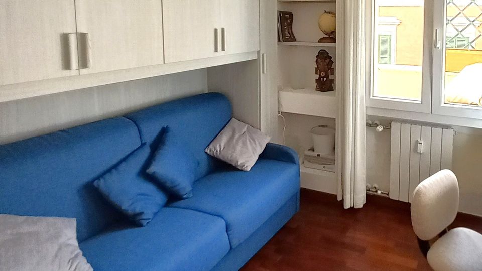 Soggiorno compatto con divano blu sotto pensili bianchi a parete, pavimento in parquet scuro e lampadario moderno. Accanto a una finestra con tende chiare e termosifone si vede una nicchia con mensole e una piccola sedia imbottita.