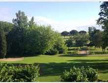 Ampio campo da golf con prato rasato e una buca in primo piano, circondato da alberi e cespugli. In lontananza si vedono ulteriori fairway e zone verdi sotto un cielo sereno.