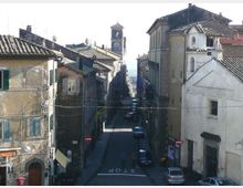 Strada stretta in un centro storico, fiancheggiata da edifici in muratura con tetti in tegole e persiane, con auto parcheggiate lungo i lati. In fondo si vede una torre con orologio che emerge sopra i tetti, mentre la via scende verso l’orizzonte.