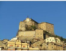Complesso fortificato in pietra su una collina, con edifici storici sovrapposti e una torre con grande orologio sulla facciata. In basso si vede il borgo con case addossate e tetti in coppi, sullo sfondo di cielo sereno.
