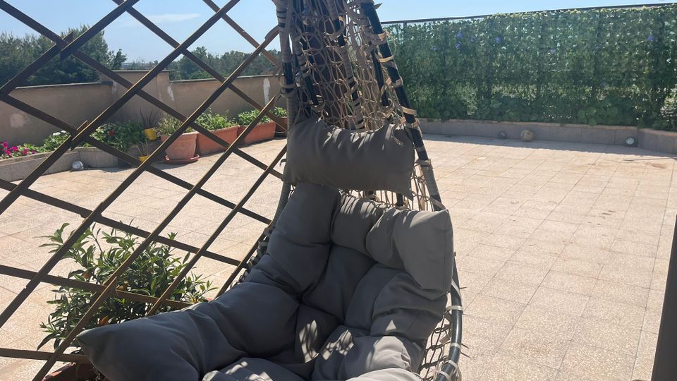 Terrazza pavimentata in piastrelle con una poltrona sospesa intrecciata e imbottita in primo piano. Sullo sfondo ci sono fioriere, una grata in legno e una recinzione coperta di vegetazione lungo il parapetto.