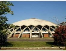 Edificio moderno a cupola, simile a un palazzetto dello sport, con grandi archi in cemento a vista lungo la facciata e ingresso centrale con vetrate. Davanti si vede una strada con prato e alberi e cespugli ai lati, sotto un cielo sereno.