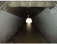 Interno di un tunnel sotterraneo con pareti e soffitto in lamiera ondulata, illuminato debolmente da una lampada centrale. In fondo si vede l’uscita circolare molto luminosa che dà sull’esterno.