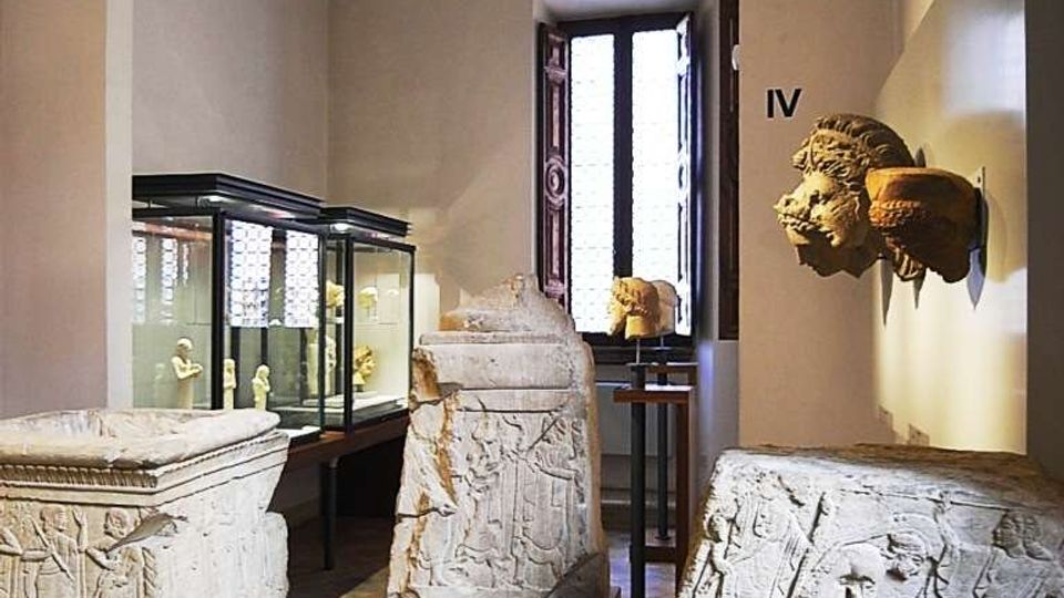Sala museale con grandi sarcofagi e frammenti lapidei scolpiti esposti su piedistalli, affiancati da vetrine in vetro illuminate. L’ambiente ha pareti chiare, un’ampia arcata interna e una finestra alta sul fondo che lascia entrare luce naturale.