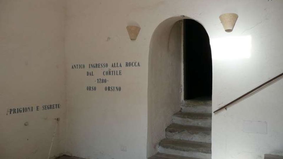 Interno di un edificio storico con pareti bianche e scritte murali; un arco conduce a una breve scalinata in pietra verso un corridoio buio. Sulla parete destra sono presenti una ringhiera e piccole applique, con luce naturale che entra da un’apertura fuori campo.