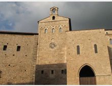 Facciata in pietra di una chiesa medievale con campanile a vela, rosone centrale e serie di archetti sotto la sommità. In basso si vede un grande portale ad arco ogivale e finestre strette, con un’ampia parete in pietra sul lato.