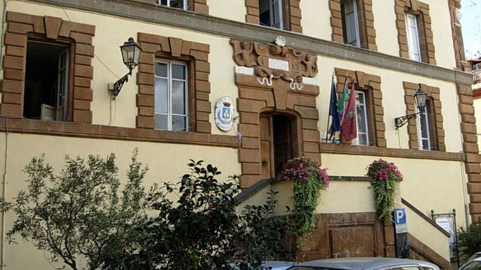 Facciata di un edificio pubblico a più piani con intonaco chiaro e cornici in pietra marrone attorno alle finestre e all’ingresso centrale sopra una scalinata. Sulla parete sono visibili uno stemma e bandiere, con fioriere ai lati e auto parcheggiate in primo piano.