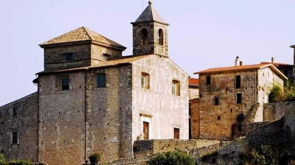 Edifici storici in pietra su un pendio, con una chiesa dal campanile quadrato e tetto a piramide in primo piano. Intorno si vedono case in muratura con tetti in coppi e un muretto di contenimento in pietra sotto un cielo limpido.