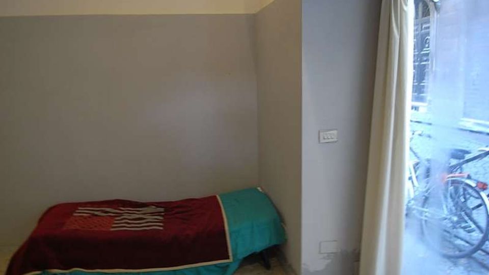 Piccola camera con letto singolo basso addossato alla parete, pareti bicolore grigio e bianco e pavimento in graniglia. A destra una grande finestra o porta-finestra con tende chiare affaccia sulla strada esterna.