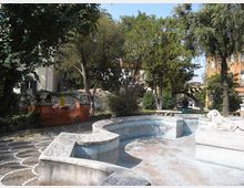 Piccolo giardino urbano con una vasca-fontana in pietra vuota dalle forme curve e basse, affiancata da un pavimento a mosaico. Alberi e cespugli circondano l’area, con edifici sullo sfondo e una recinzione arancione da cantiere su un lato.