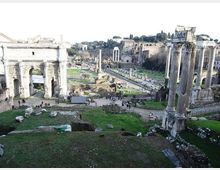 Veduta panoramica del Foro Romano con rovine antiche, includendo un grande arco in pietra sulla sinistra e alte colonne di un tempio sulla destra. Al centro si estende l’area archeologica con resti di edifici e percorsi tra prati e pietre, sullo sfondo delle colline e della città.