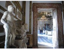 Interno di una galleria museale con statue in marmo lungo le pareti, cornici decorative e una grande apertura incorniciata in marmo che conduce a una sala ampia con pavimento a scacchi e lampadari. Sullo sfondo si vedono ulteriori sculture esposte su piedistalli in un ambiente illuminato da luce naturale.