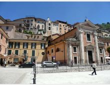 Piazza urbana con una chiesa in stile classico in primo piano, con facciata in mattoni e pietra, portale centrale e timpano sormontato da una croce. Sullo sfondo si vedono edifici residenziali dai colori chiari disposti su un pendio, con un campanile e cielo azzurro sopra.