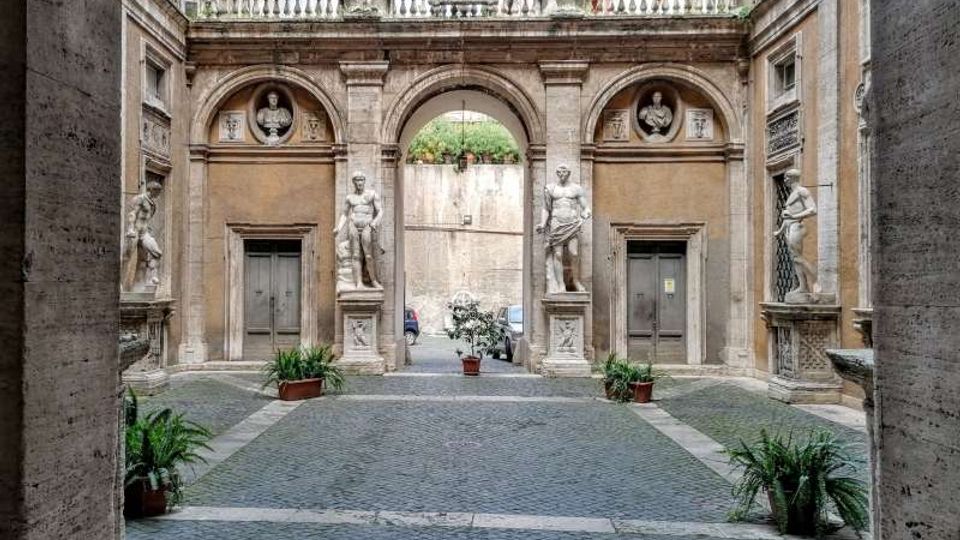 Cortile in pietra con pavimentazione a sampietrini, delimitato da una facciata classica con tre archi e balconata superiore. Ai lati dell’arco centrale si vedono statue in nicchie e due portali simmetrici, con vasi di piante disposti lungo il passaggio.