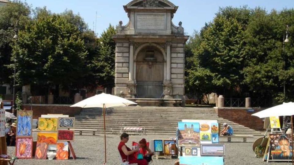 Piazza lastricata con grande scalinata che conduce a un monumento in pietra con nicchia ad arco e frontone decorato, circondato da alberi. In primo piano sono visibili cavalletti e pannelli con dipinti esposti sotto ombrelloni, tipici di un mercato d’arte all’aperto.
