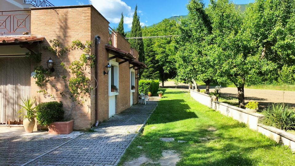 Casa in mattoni con portico e finestre affacciate su un vialetto pavimentato e un prato verde. A destra si vede un frutteto con alberi e un basso muretto, con colline boscose sullo sfondo sotto un cielo azzurro.