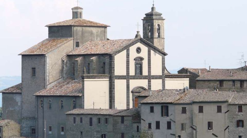 Chiesa in muratura su un borgo collinare, con facciata chiara incorniciata da pietra scura e campanile quadrato con aperture ad arco. Intorno si vedono edifici addossati con tetti in coppi e poche finestre, sullo sfondo cielo chiaro.