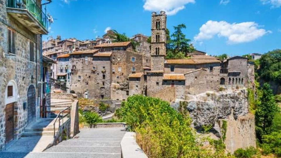Borgo storico in pietra su uno sperone roccioso, con case addossate e una torre campanaria che domina il centro abitato. In primo piano una passerella con ringhiera e scalinate conduce verso il paese, affiancata da vegetazione e alberi sotto un cielo azzurro.