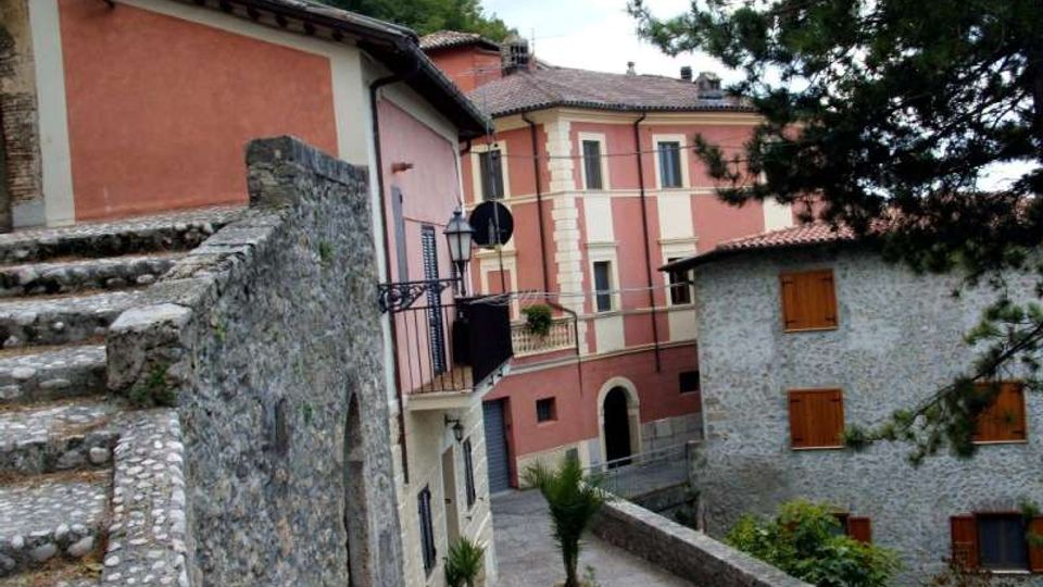 Vicolo in un borgo collinare con una scalinata in pietra e un muretto che conduce a case ravvicinate. Gli edifici includono una casa intonacata rosa con balconcino e un edificio in pietra con persiane in legno, circondati da alberi e vegetazione.