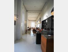 Interno di un ristorante o caffetteria in stile moderno, con lungo corridoio, pavimento chiaro lucido e soffitto alto. A destra si vede un bancone con vetrina e scaffali con bottiglie, mentre in fondo ci sono tavoli con sedie e una grande finestra che illumina la sala.