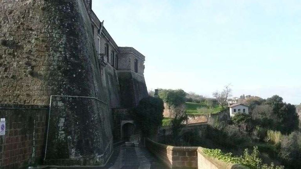 Camminamento lastricato lungo una grande fortificazione in pietra con torre e mura massicce, con un passaggio ad arco in fondo. A destra un muretto di mattoni affaccia su una valle verde con alberi e alcune case in lontananza.