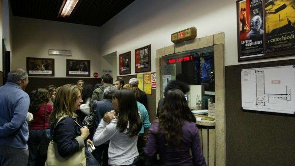 Interno di un cinema con folla in coda davanti alla biglietteria incassata nel muro, con display luminoso sopra lo sportello. Sulle pareti sono affissi poster di film e a destra è visibile una piantina dell’edificio su pannello.