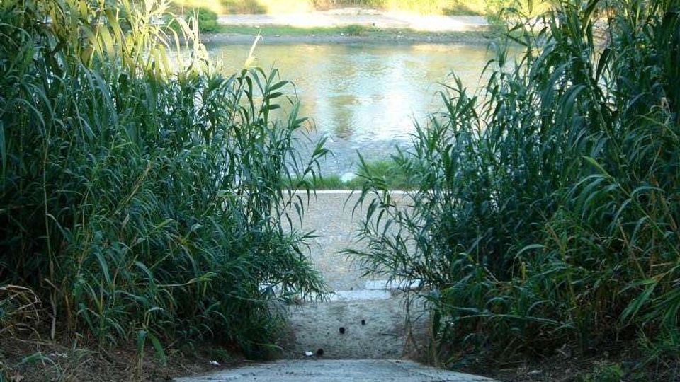 Scalinata in cemento che scende tra canneti e vegetazione fitta fino alla riva di un fiume o canale. Sullo sfondo si vede l’acqua e la sponda opposta con erba e alberi.