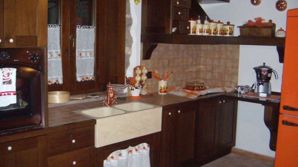 Cucina domestica con mobili in legno scuro a L, piano di lavoro con lavello e rubinetto in rame e alzatina piastrellata. Si vedono finestra con tende, forno incassato, mensole con barattoli e utensili, pavimento in piastrelle e un frigorifero arancione sulla destra.