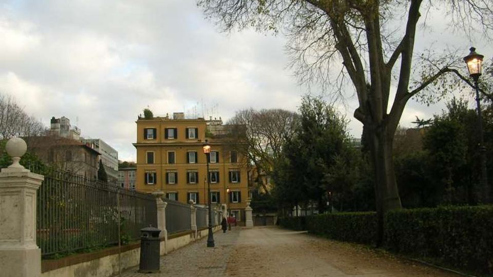Viale pedonale in un parco urbano con percorso in ghiaia, siepi e grandi alberi spogli ai lati. A sinistra ci sono una recinzione in ferro con muretto e lampioni, sullo sfondo un edificio giallo a più piani sotto cielo nuvoloso.