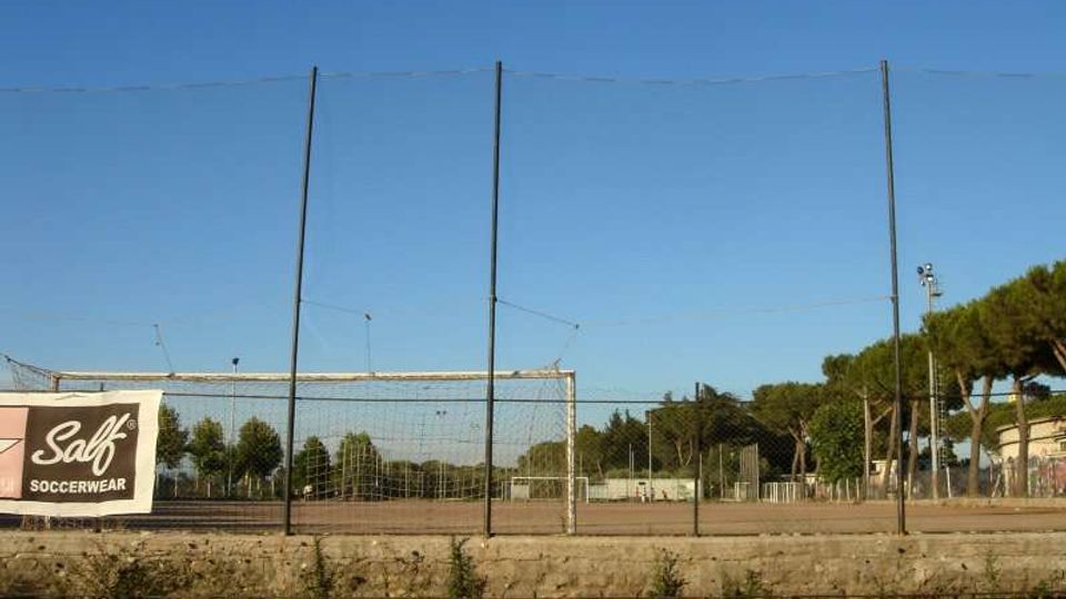 Campo sportivo all’aperto recintato con rete alta e pali metallici, visto da un parcheggio con strisce bianche. Sullo sfondo si notano alberi, un palo faro e un cartellone pubblicitario sul lato sinistro sotto un cielo sereno.