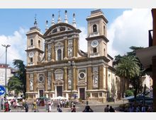 Facciata di una grande chiesa in stile barocco con due campanili laterali dotati di orologi, statue in nicchie e frontone decorato con croci e pinnacoli. Davanti si apre una piazza con gradinate, bancarelle e alberi, circondata da edifici urbani.