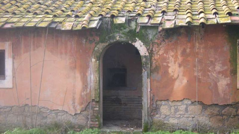 Facciata di un edificio rurale con intonaco scrostato e tetto in coppi, con un arco centrale che conduce a un ingresso buio con gradini interni. Alla base si vede terreno erboso con vegetazione bassa e tracce di umidità e muschio attorno all’arco.