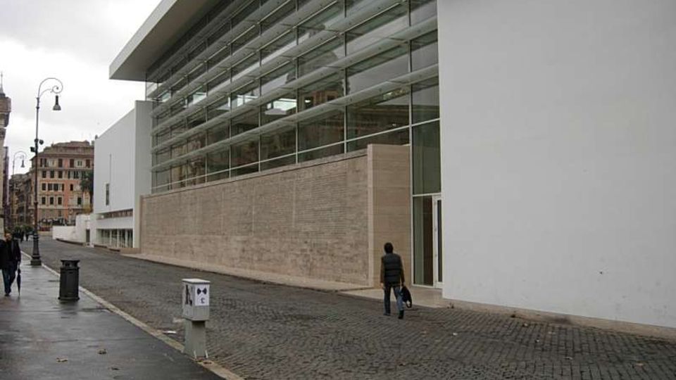 Edificio moderno con lunga facciata vetrata a più livelli e ampie pareti bianche, affacciato su una strada in pavé. L’ingresso è vetrato e sullo sfondo si vedono lampioni e palazzi urbani.