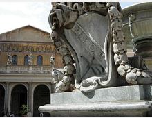 Dettaglio ravvicinato di una grande fontana in pietra scolpita, con elementi decorativi curvilinei e una vasca circolare sul lato destro. Sullo sfondo si vede un edificio storico con portico ad archi, statue e facciata con mosaici.