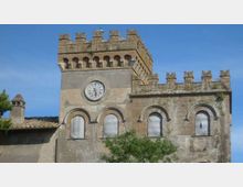 Edificio storico in pietra con torre merlata e orologio circolare sulla facciata, affiancato da un corpo con finestre ad arco e parapetto merlato. Sullo sfondo cielo azzurro, con tetti in coppi e vegetazione in primo piano.
