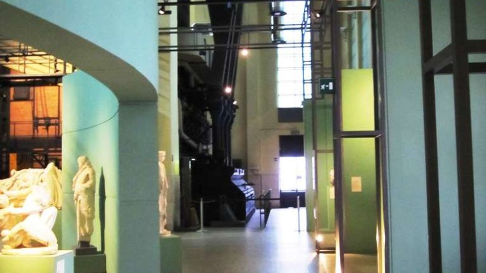 Interno di un museo in un ampio corridoio con pareti verde acqua, pavimento a mosaico chiaro e grandi condotte metalliche a vista sul soffitto. Lungo il percorso si vedono teche e piedistalli con statue, strutture metalliche verticali e un’alta finestra sul fondo che illumina lo spazio.