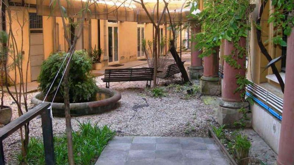 Cortile interno di un edificio con pavimentazione in ghiaia e un breve tratto di piastrelle, aiuole e alberi; al centro si vedono panchine e una grande fioriera circolare. Sui lati ci sono facciate con porte e finestre e un porticato con colonne cilindriche rosa.
