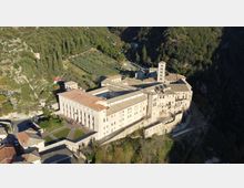 Veduta aerea di un grande complesso monastico in pietra con tetti in coppi e un campanile quadrato, disposto attorno a cortili e terrazze su un pendio. L’edificio è circondato da colline boscose e campi coltivati, con strade e piccoli edifici rurali nelle vicinanze.