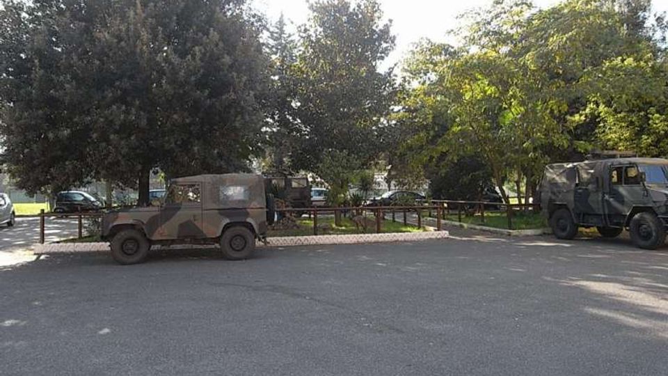 Parcheggio asfaltato ai margini di un’area verde con alberi alti e recinzione in legno. Lungo il bordo sono presenti diversi veicoli fuoristrada, alcuni con livrea mimetica, vicino a una zona erbosa.