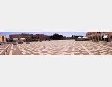 Ampia piazza pedonale pavimentata con motivi geometrici a riquadri chiari e scuri, con dissuasori in cemento e lampioni. Sullo sfondo si vedono edifici moderni in mattoni rossi, alcune aiuole e una struttura coperta con teli bianchi.