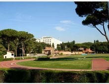 Campo sportivo all’aperto con pista di atletica rossa che circonda un’ampia area erbosa. Sullo sfondo si vedono pini mediterranei, piccoli edifici e una grande struttura bianca multipiano sotto un cielo sereno.