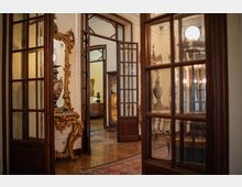 Interno di un palazzo storico con porte a vetri e telai in legno scuro che si aprono su stanze comunicanti. Si vedono pavimento in parquet, tappeti e arredi d’epoca con specchi e consolle intagliate e vasi decorativi.