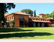 Edificio in stile villa mediterranea con facciata color terracotta e tetti in coppi, caratterizzato da grandi finestre ad arco e porticati con colonne. Davanti si estende un ampio prato curato, con alberi alti e vegetazione sullo sfondo.