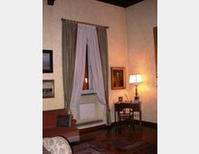 Interno di una sala in stile classico con pareti chiare, pavimento in legno e finestra alta con tende drappeggiate. Accanto alla finestra ci sono un piccolo scrittoio con lampada da tavolo e cornici, quadri appesi alle pareti, un divano e tappeti decorati.