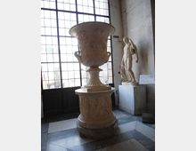 Sala museale con grande finestra a griglia e pavimento in marmo; al centro un grande cratere in pietra decorato su piedistallo, con una statua classica su basamento lungo la parete.
